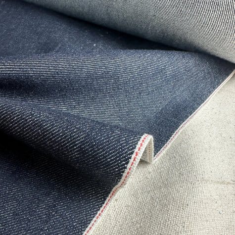 Close-up of Japanese selvedge denim fabric showing dark indigo twill texture and a clean self-finished edge with red stitching.