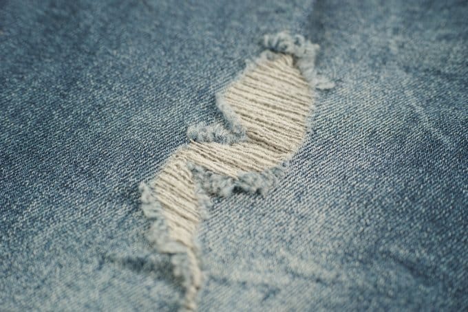 Close-up of distressed washed denim with a torn hole and frayed threads on light blue fabric.