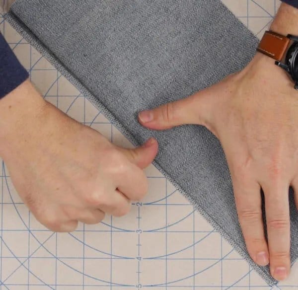 Hands folding and preparing chambray fabric for hemming on a cutting mat, demonstrating garment finishing techniques.