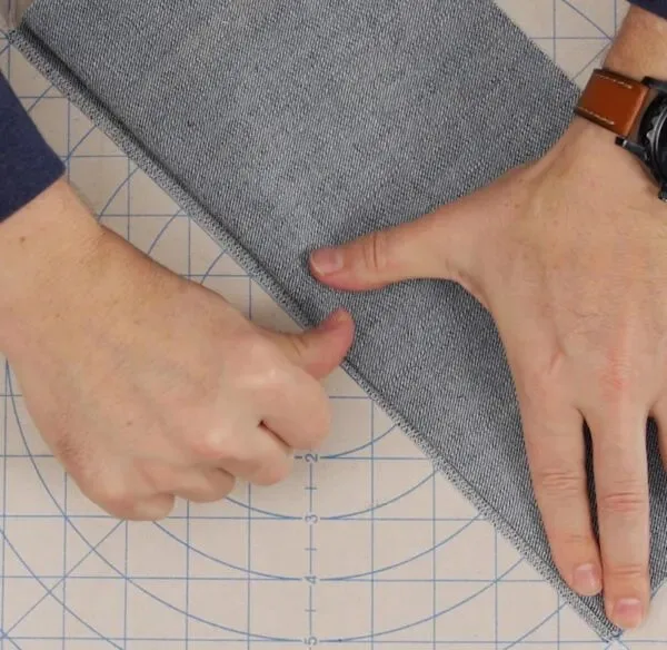 Hands folding and preparing chambray fabric for hemming on a cutting mat, demonstrating garment finishing techniques.