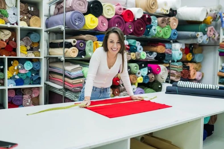Fabric supplier measuring material on a cutting table surrounded by colourful textile rolls in a fabric warehouse.