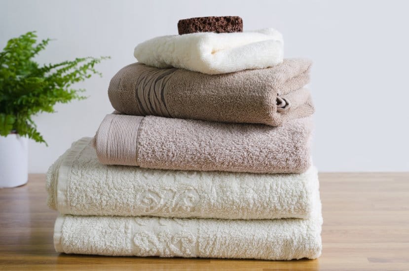 Stack of soft cream, beige and brown cotton bath towels arranged on a wooden surface beside a green plant.