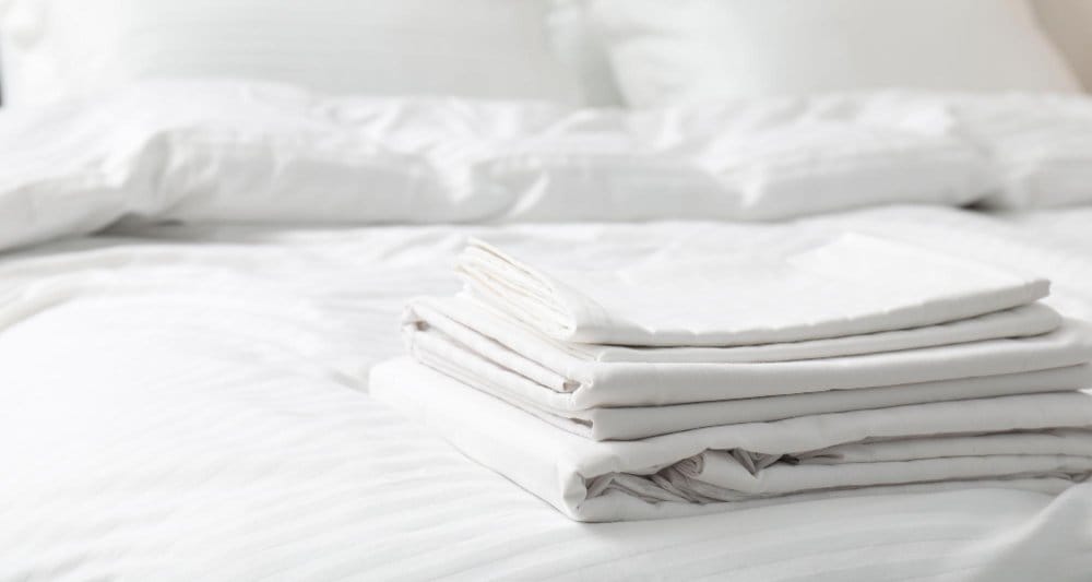 Stack of clean white cotton bed linens arranged on a neatly made bed with white pillows and bedding.