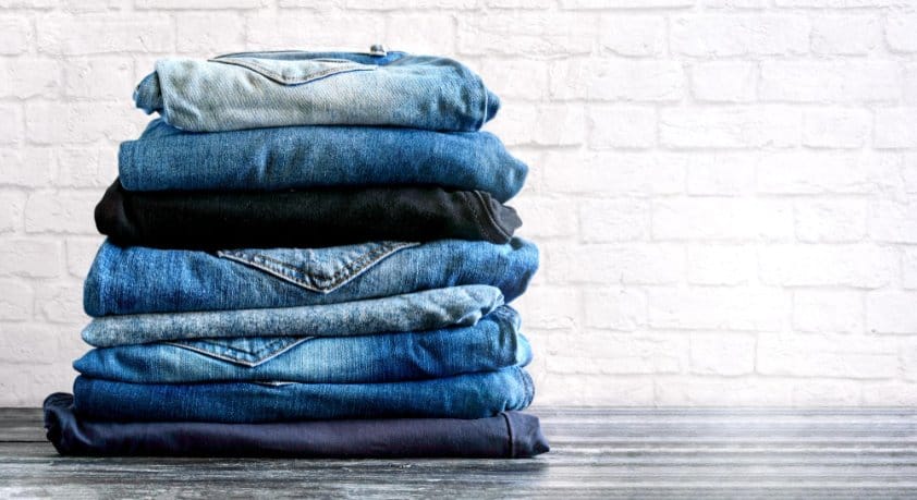Stack of folded blue and black denim jeans in different washes arranged against a white brick wall.