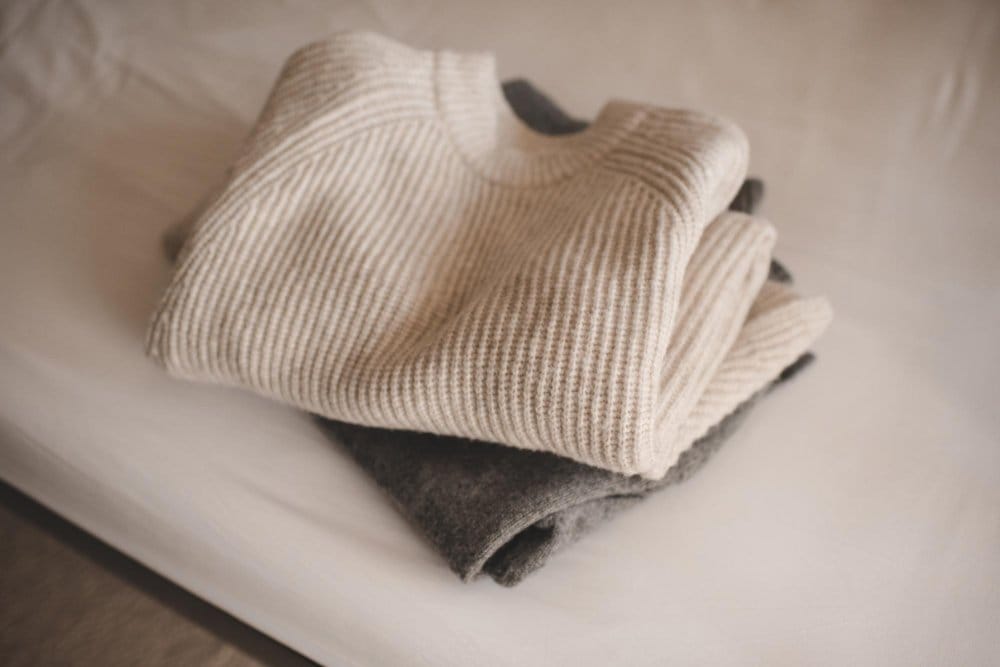 Neatly folded cream and grey knitted sweaters stacked on a bed, showing the soft texture of fine knitwear.