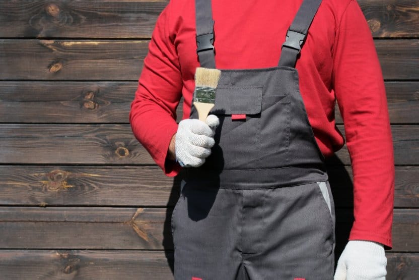 Person wearing grey workwear overalls, a red long-sleeved top and protective gloves while holding a paintbrush.